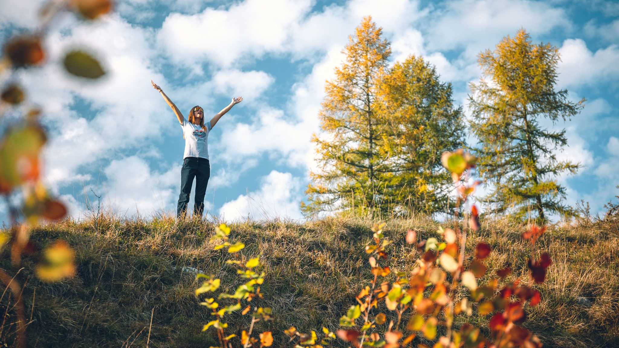 Eine Frau steht auf einer Wiese mit erhobenen Armen vor blauem Himmel und weißen Wolken; neben ihr leuchten herbstlich gelbe Bäume, im Vordergrund unscharfe Blätter.