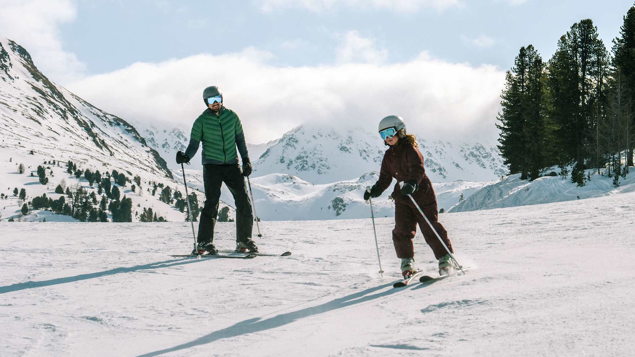 Ein Erwachsener und ein Kind fahren zusammen Ski auf einer sonnigen Piste in Nauders, umgeben von verschneiten Bergen.