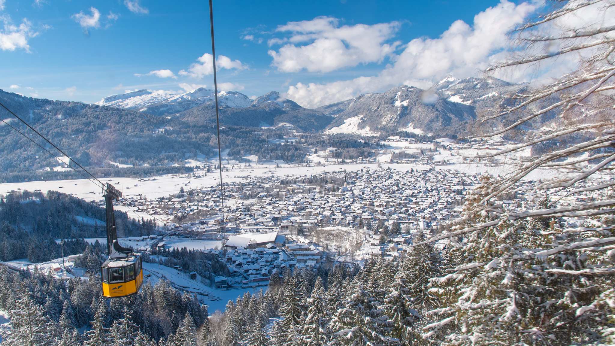 Blick von oben auf die gelbe Nebelhornbahn mit eingeschneiten Häusern und Bergen im Hintergrund