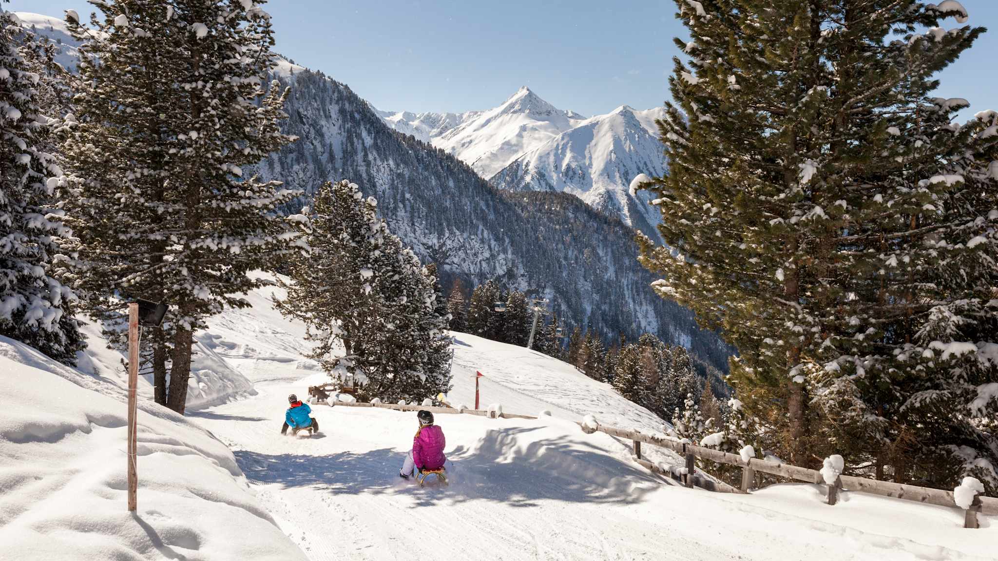 Zwei Kinder rodeln am Hochzeiger im Pitztal, Tirol, auf einem verschneiten Waldweg mit Blick auf die umliegenden Berge.