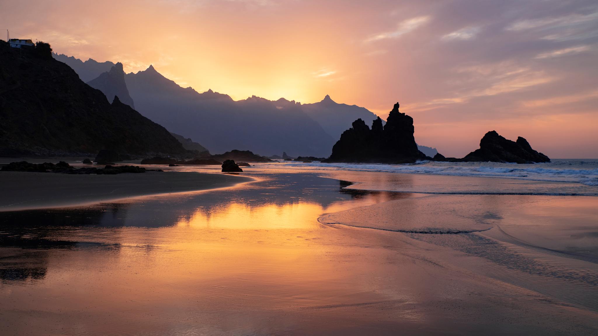Sonnenuntergang am menschenleeren Sandstrand. In der Ferne sind Felsen und Gebirge zu sehen.