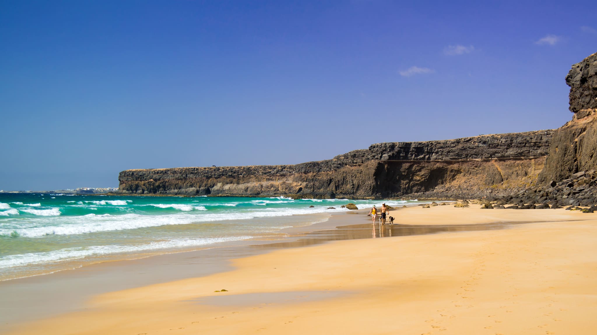 Blick auf die Naturstrand-Bucht Playa del Águila, Fuerteventura mit Steilküste und zwei Menschen mit Hund am Strand