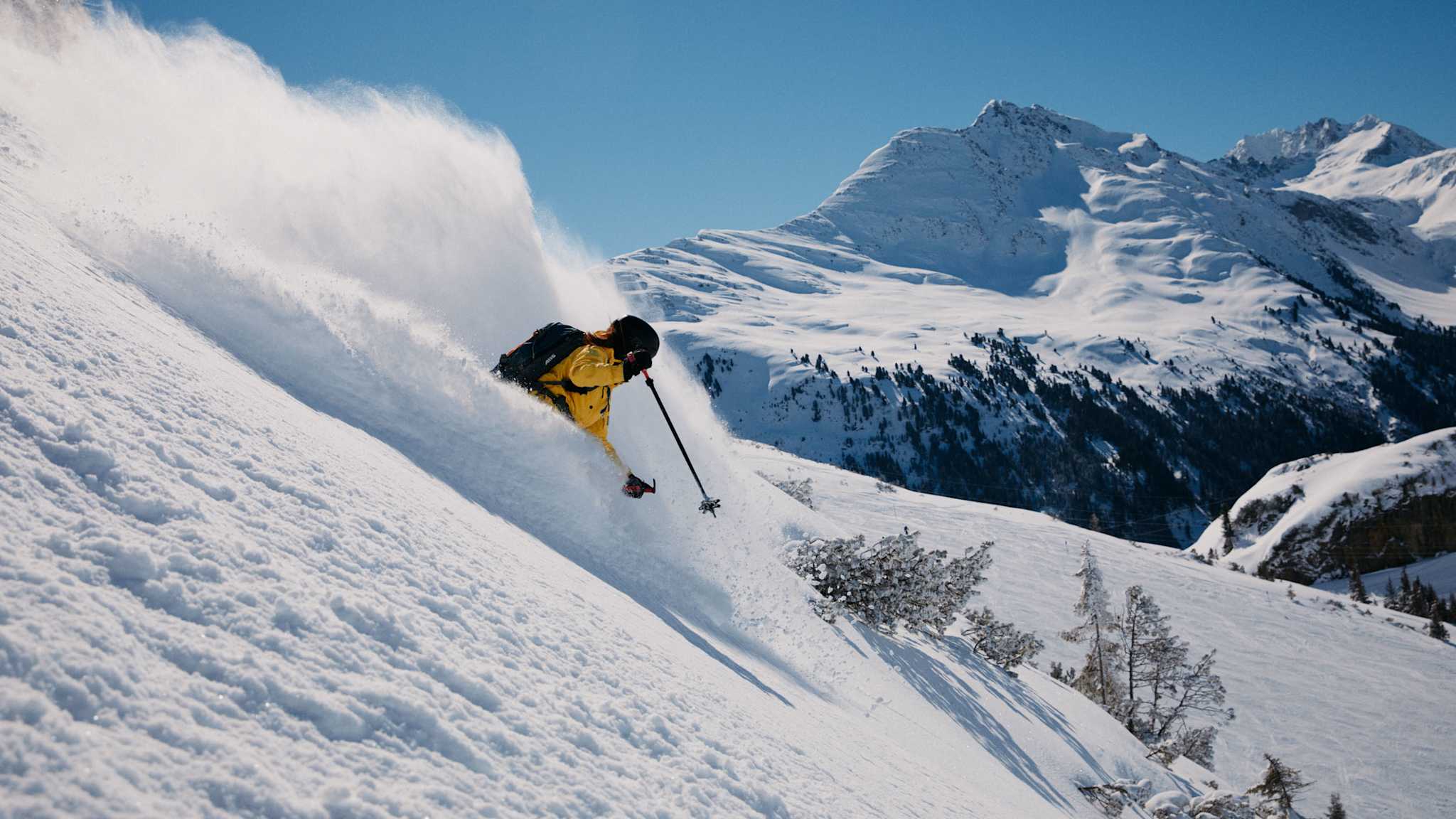 Eine Person rast auf Skiern einen Hang hinab. Dabei wirbelt Schnee auf.