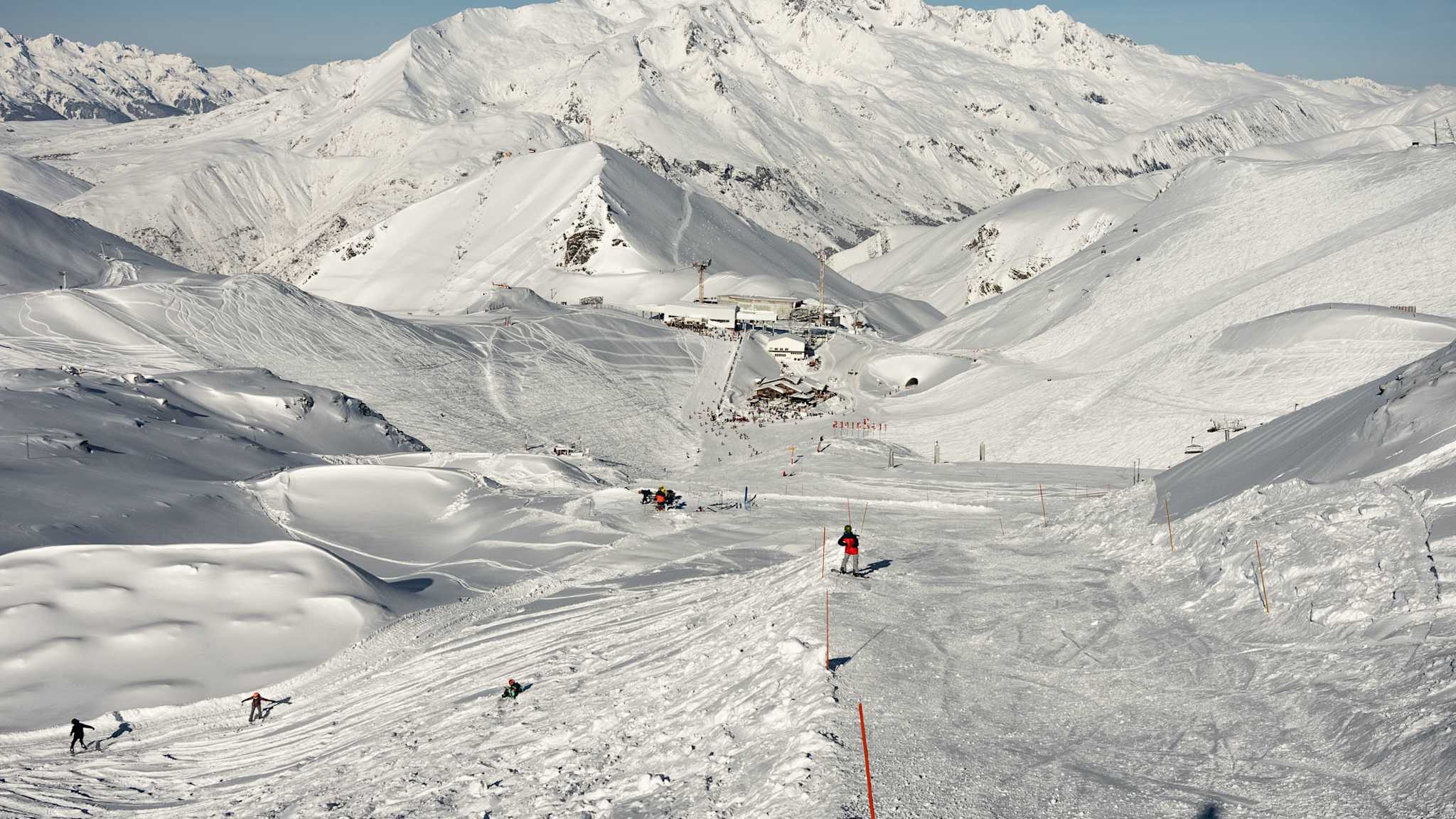 Skifahrer auf einer breiten, sonnigen Skipiste mit umliegenden weißen Berggipfeln im Wintersportgebiet Les Deux Alpes in Frankreich.