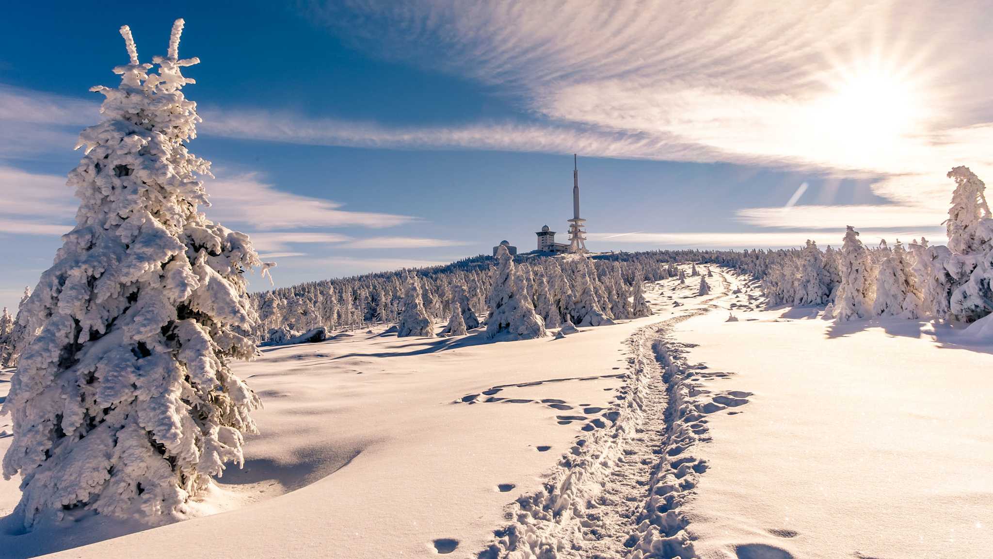Ein verschneiter Wanderweg führt durch eine weiße Winterlandschaft auf den Brocken im Harz, im Hintergrund der Sendeturm.