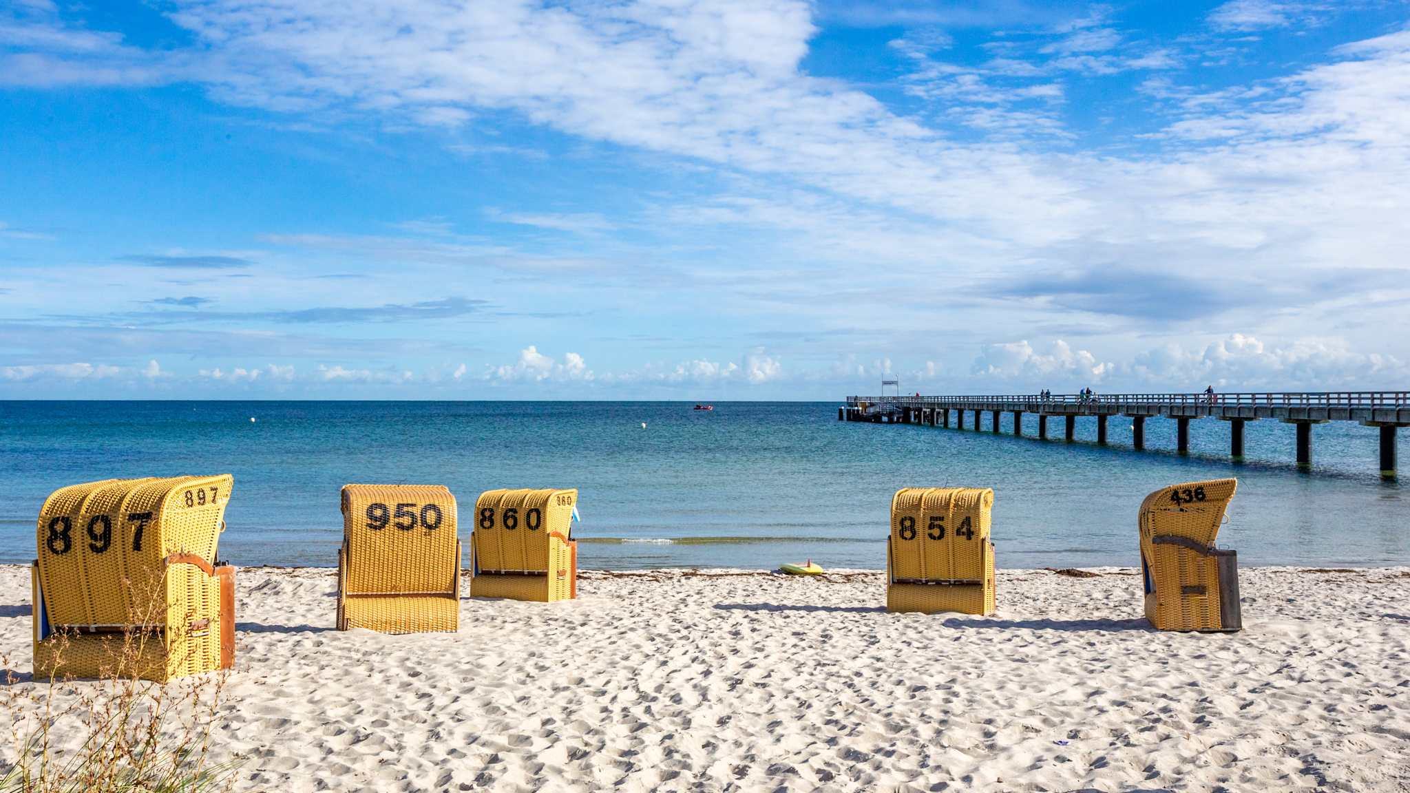 Blick über einen weichen Sandstrand auf das glatte Meer. Auf dem Sand stehen gelbe Strandkörbe. Im Hintergrund führt eine Seebrücke aufs Meer.