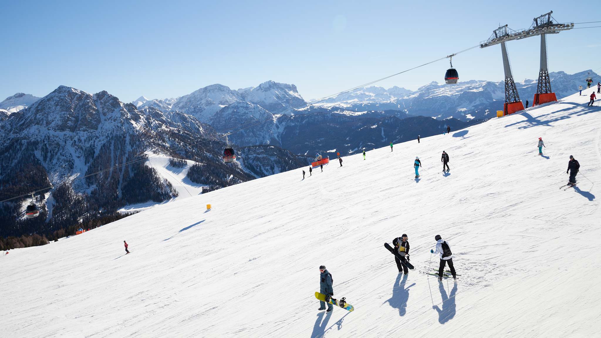 Blick auf eine sonnige Skipiste mit Skifahrenden und schneebedeckten Bergen im Hintergrund
