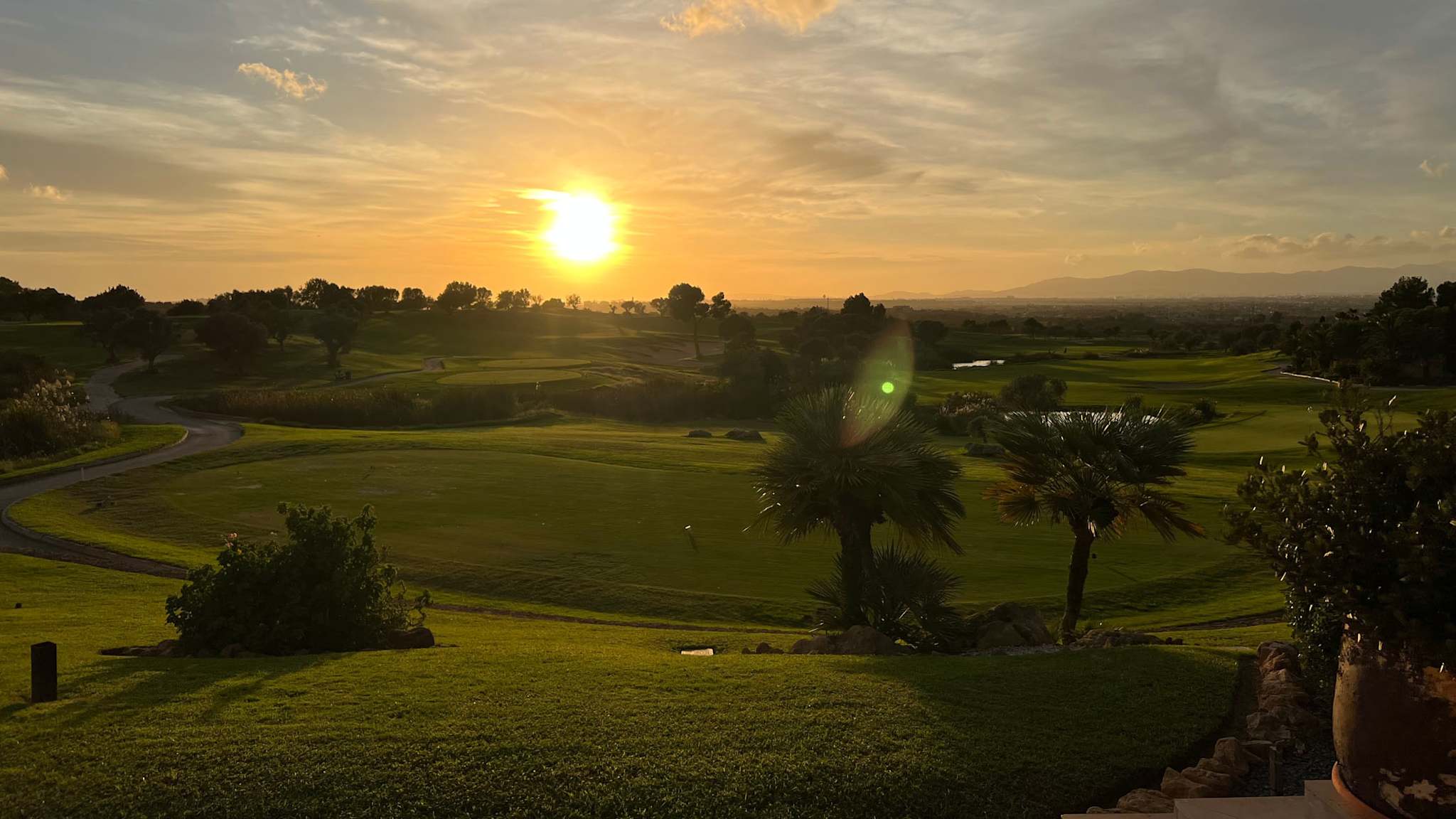 Ein weitläufiger, gepflegter Golfplatz mit Palmen und hügeligem Grün wird in warmes Licht eines Sonnenuntergangs getaucht, im Hintergrund Berge und weite Landschaft bei Palma de Mallorca.