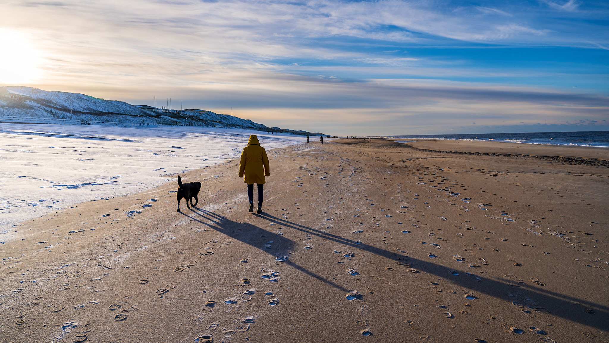 Frau im gelben Mantel geht mit Hund am verschneiten Strand von Sylt entlang, im Hintergrund die Nordsee.