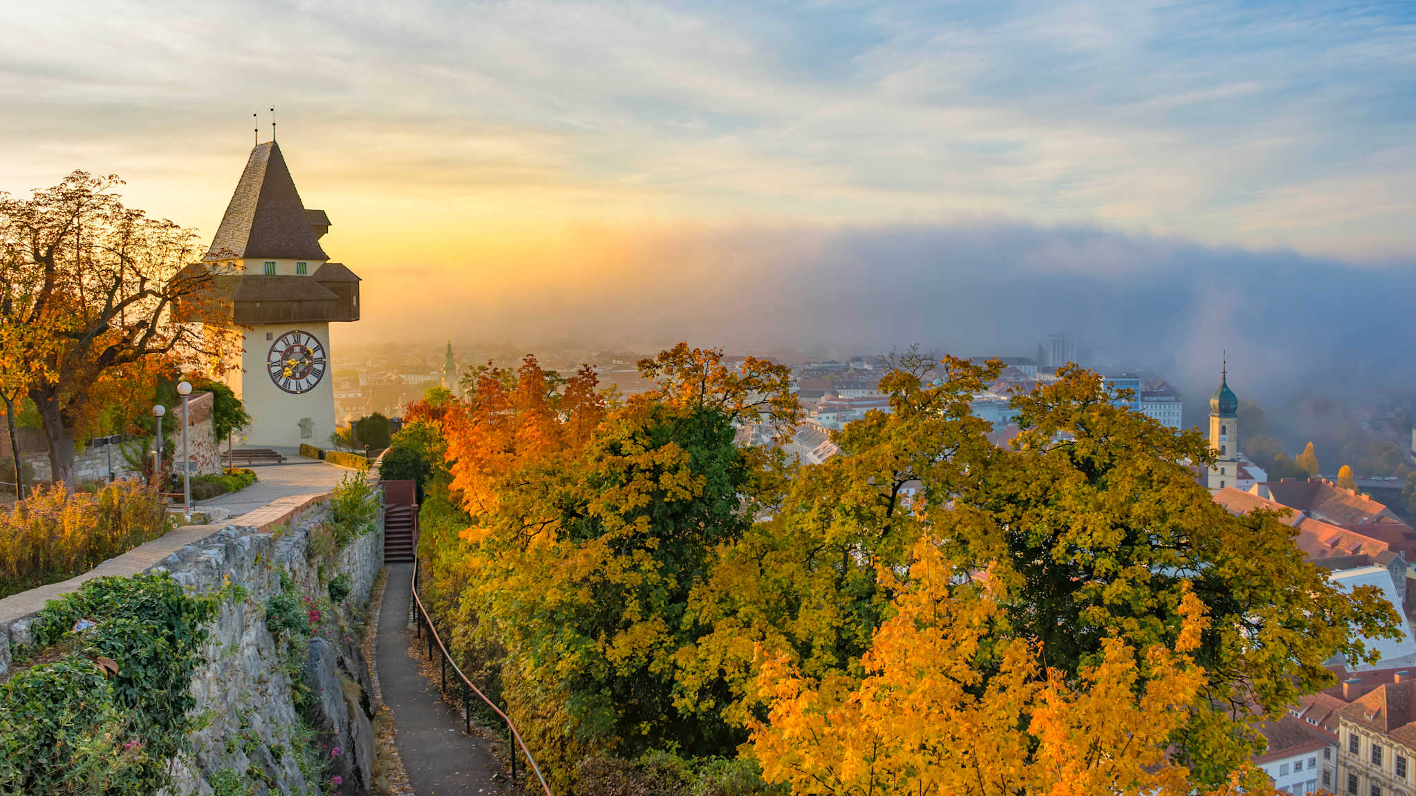 Ausblick auf den Uhrturm auf dem Schlossberg in Granz auf herbstfarbene Bäume vor der tiefer liegenden Stadt Grad, die teilweise von Nebelschwaden bedeckt ist.