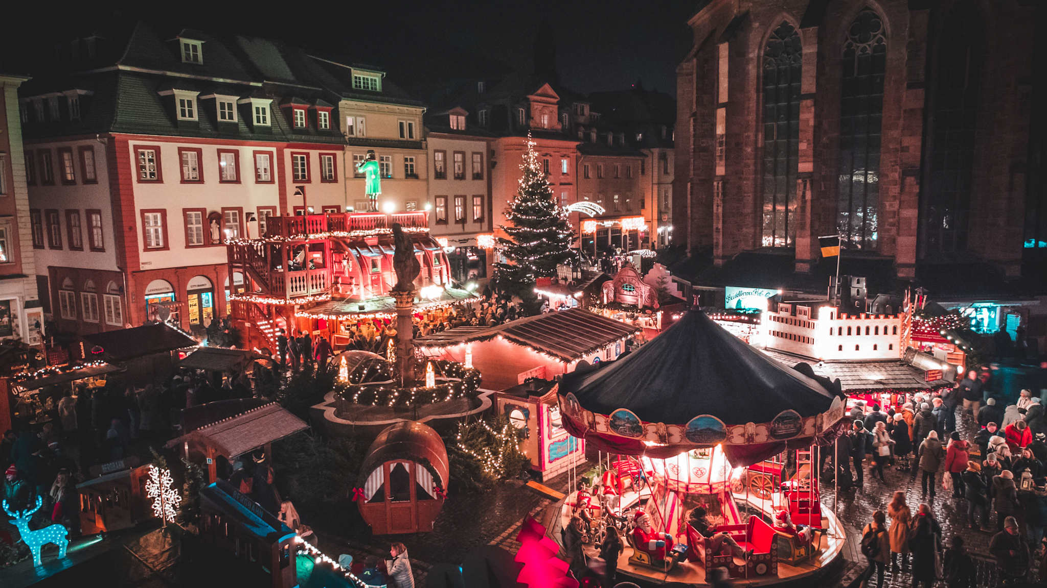 Blick auf den festlich beleuchteten Weihnachtsmarkt in Heidelberg bei Nacht, mit Karussell, Marktständen, einem Weihnachtsbaum und den umliegenden historischen Gebäuden sowie der großen Kirchenfassade im Hintergrund.