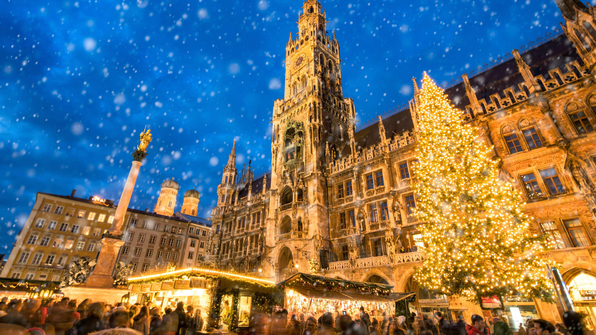 Winterliche abendliche Szene des Christkindlmarkts am Marienplatz in München, mit dem beleuchteten Rathaus, einem großen geschmückten Weihnachtsbaum und Schneefall über den Marktständen.