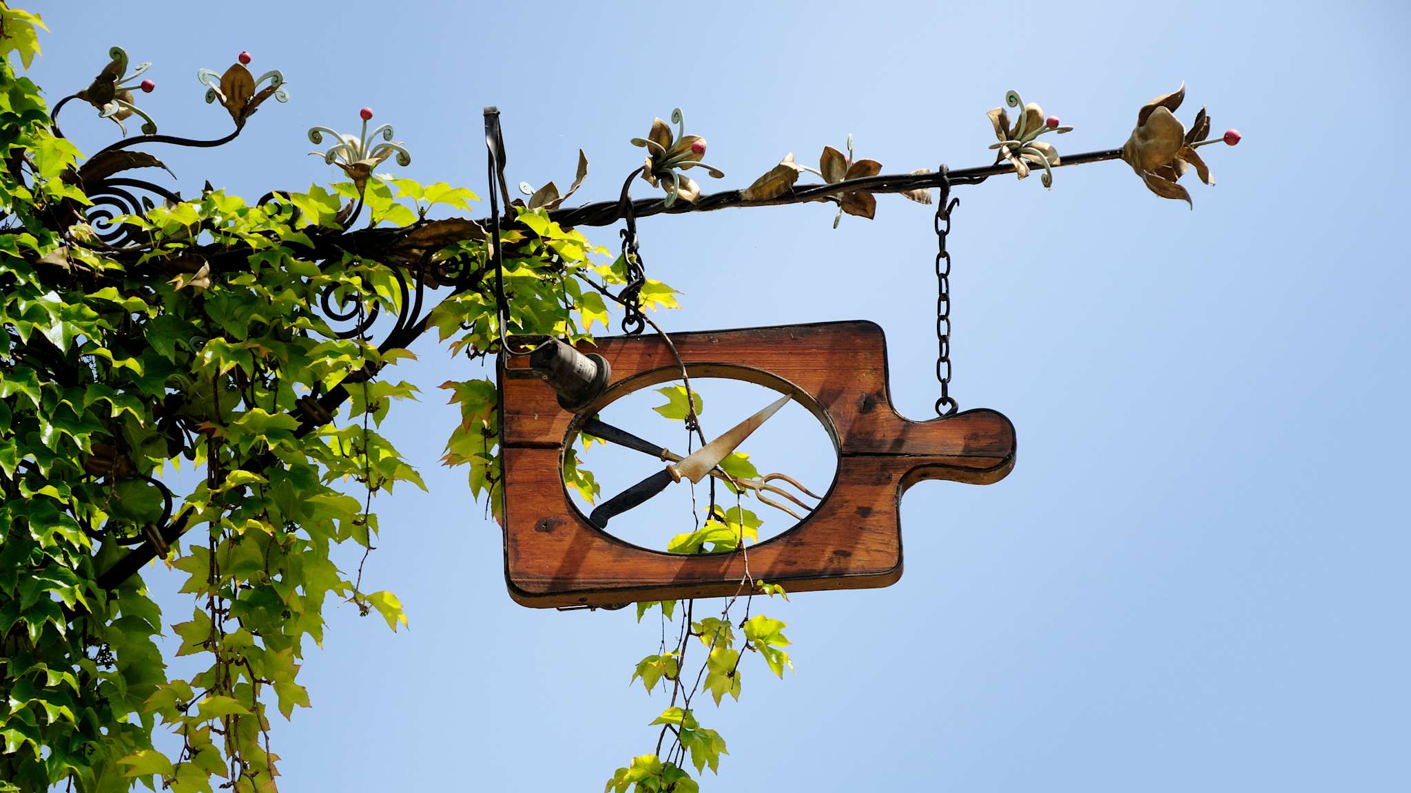 Ein Holschild in Form eines Holz-Schneidebretts mit Messer und Gabel überkreuzt in der Mitte. Es hängt an einem mit Blumen verzierten Metallgiebel, der von Efeu berankt ist, vor blauem Himmel.