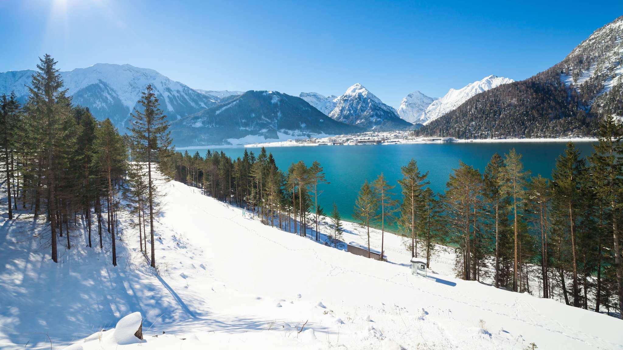 Sonnige Winterlandschaft in Tirol mit schneebedeckten Bergen, Nadelwald und türkisblauem Achensee.