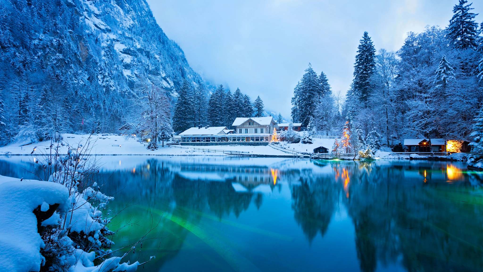 Verschneite Winterlandschaft am Blausee im Berner Oberland, mit klarem, blauem Wasser, umgeben von Wald und einem beleuchteten Haus am Ufer.