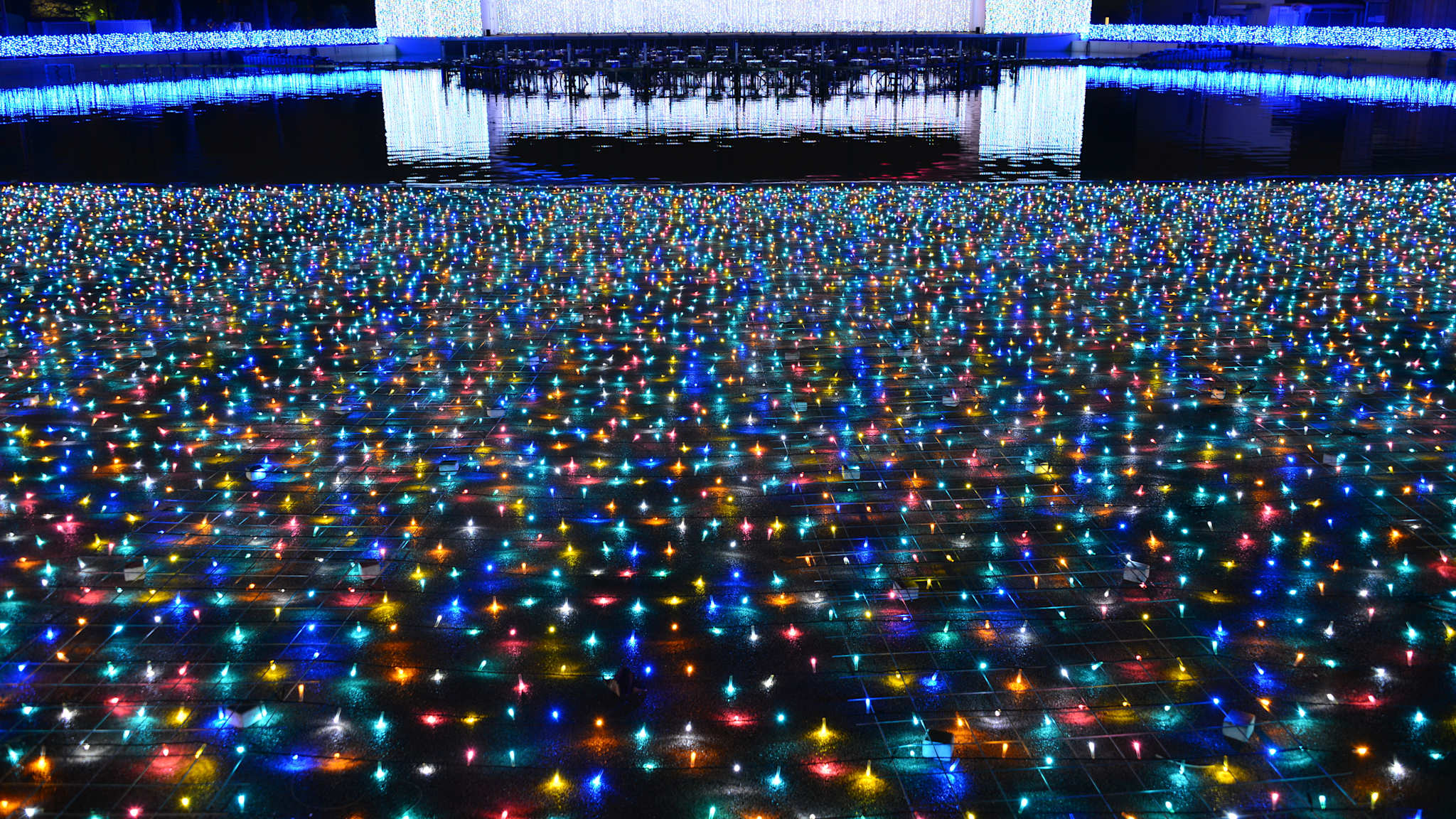 Buntes Netz aus Lichterketten auf dem Boden vor dem Wasser mit beleuchteter Architektur am Ufer.
