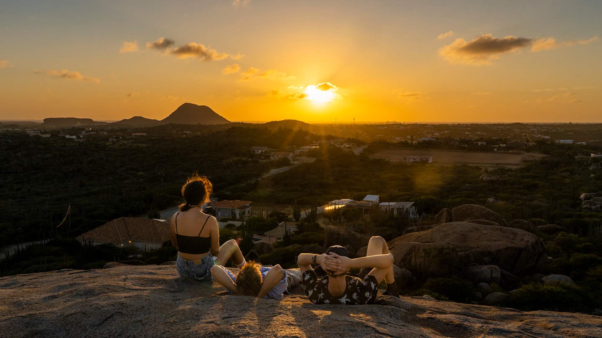 Drei junge Leute sitzen auf einem Felsen und schauen über eine Ebene auf den Sonnenuntergang.