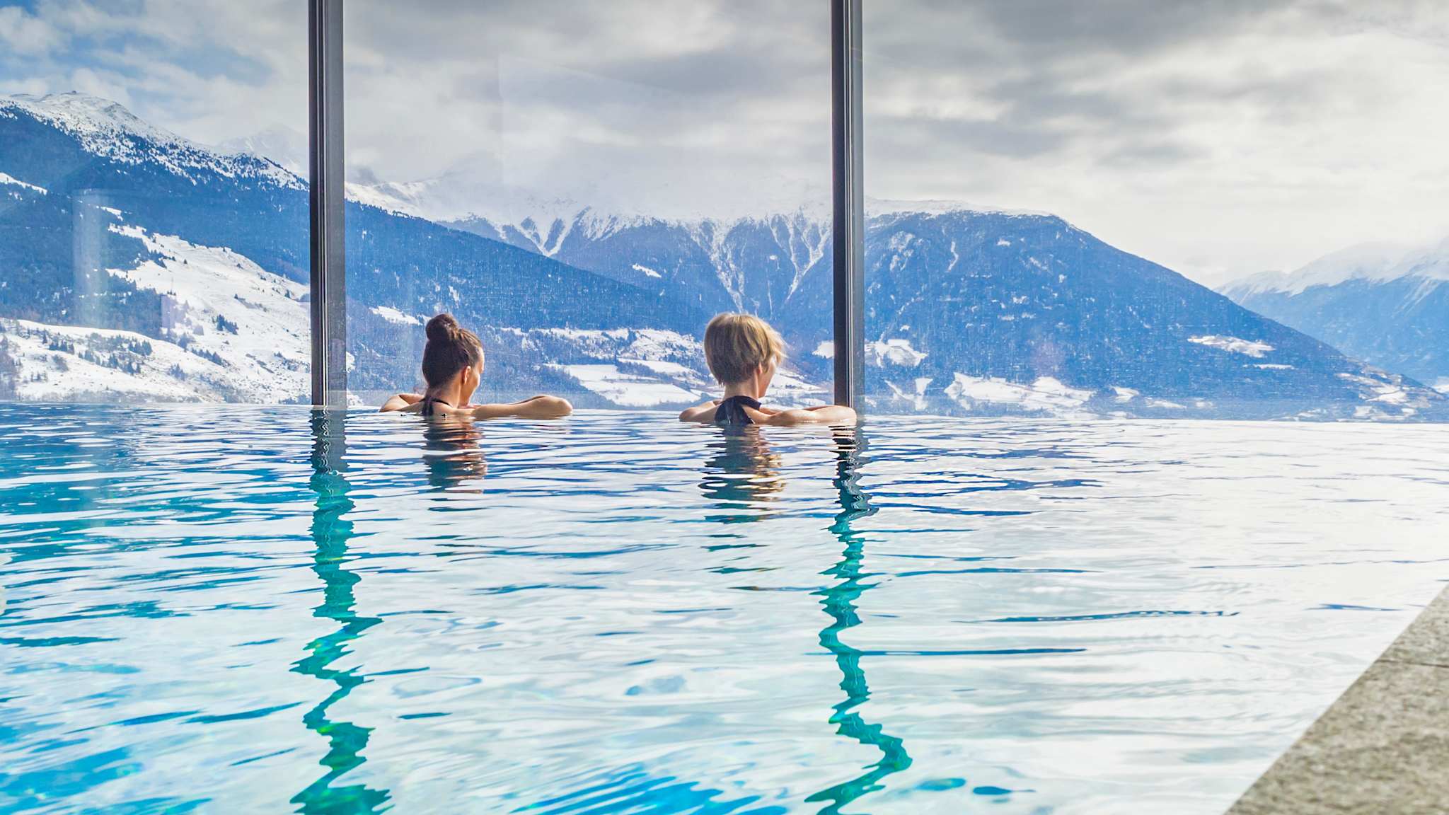 Zwei Frauen in einem Pool blicken auf eine bergige Landschaft