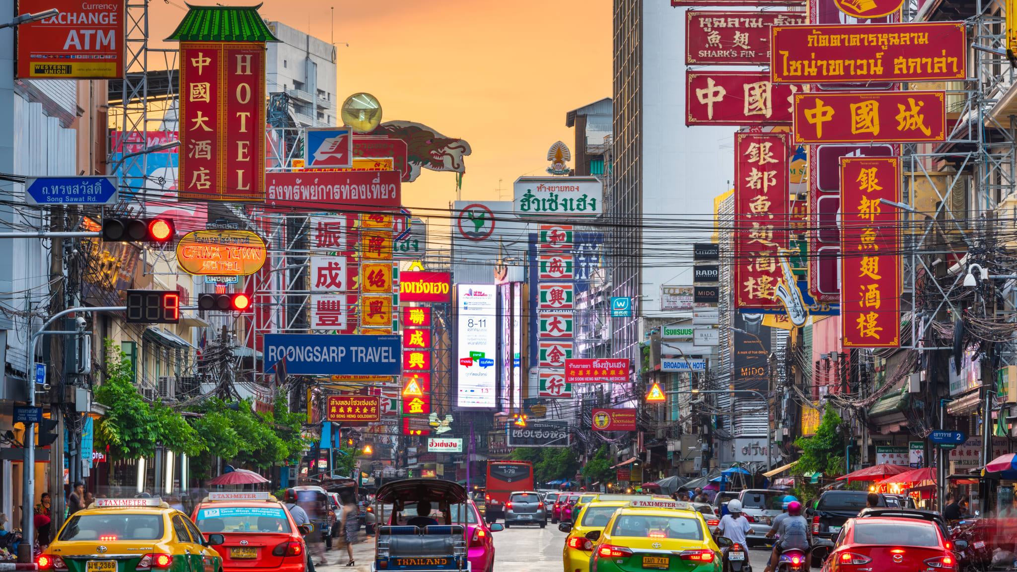 Autos fahren zwischen Hochhäusern entlang. Überall sind Werbeschilder mit thailändischer, chinesischer und englischer BEschriftung zu sehen.