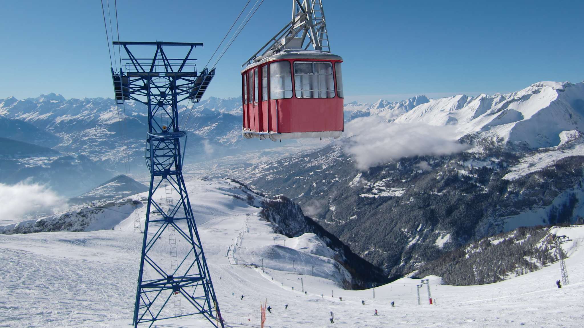 Die Gondel einer Seilbahn schwebt über einer Skipiste vor einer Alpenkulisse.