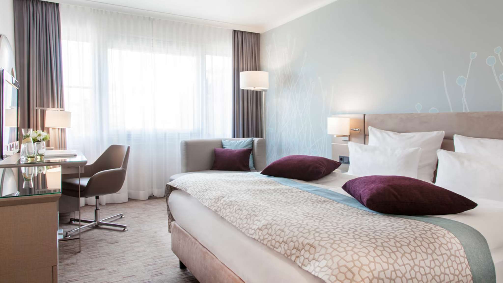 Ein Doppelzimmer in einem Hotel ist eingerichtet mit einem Schreibtisch, einem Sessel und einem Doppelbett.