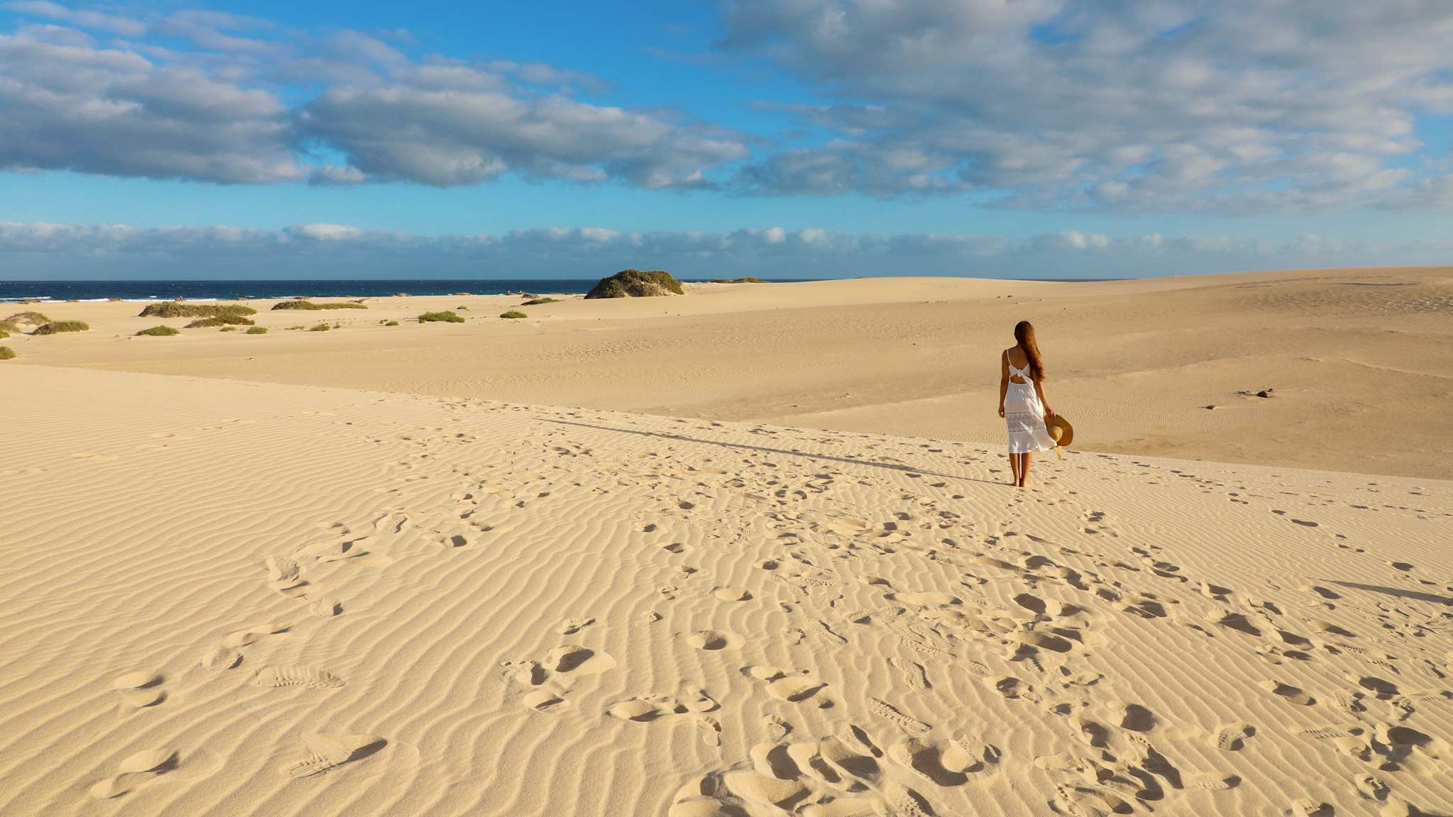 Eine Frau läuft im weißen Sommerkleid und mit Hut in der Hand über die Dünen bei Corralejo, Fuerteventura