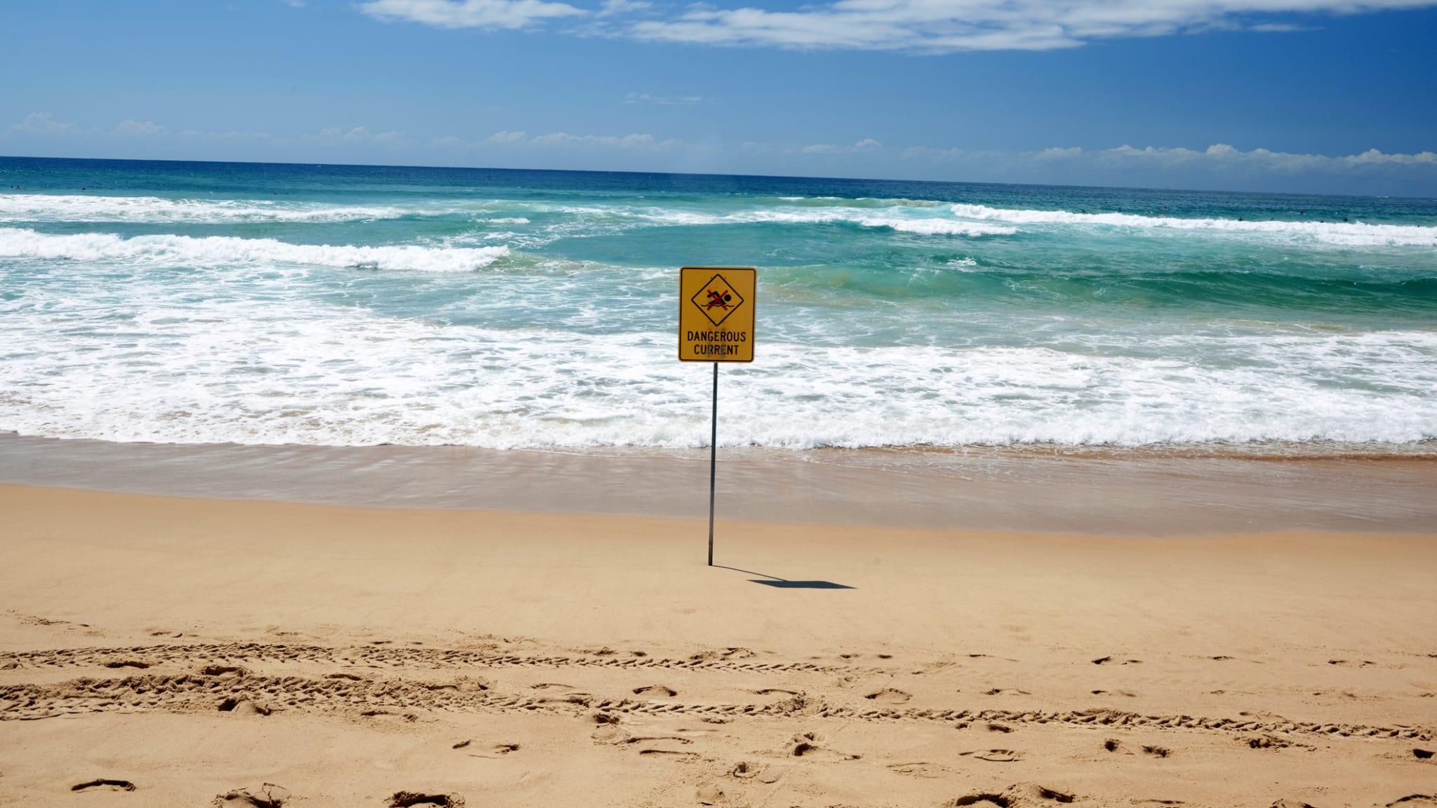 Ein gelbes Warnschild an einem Strand direkt am Meer.