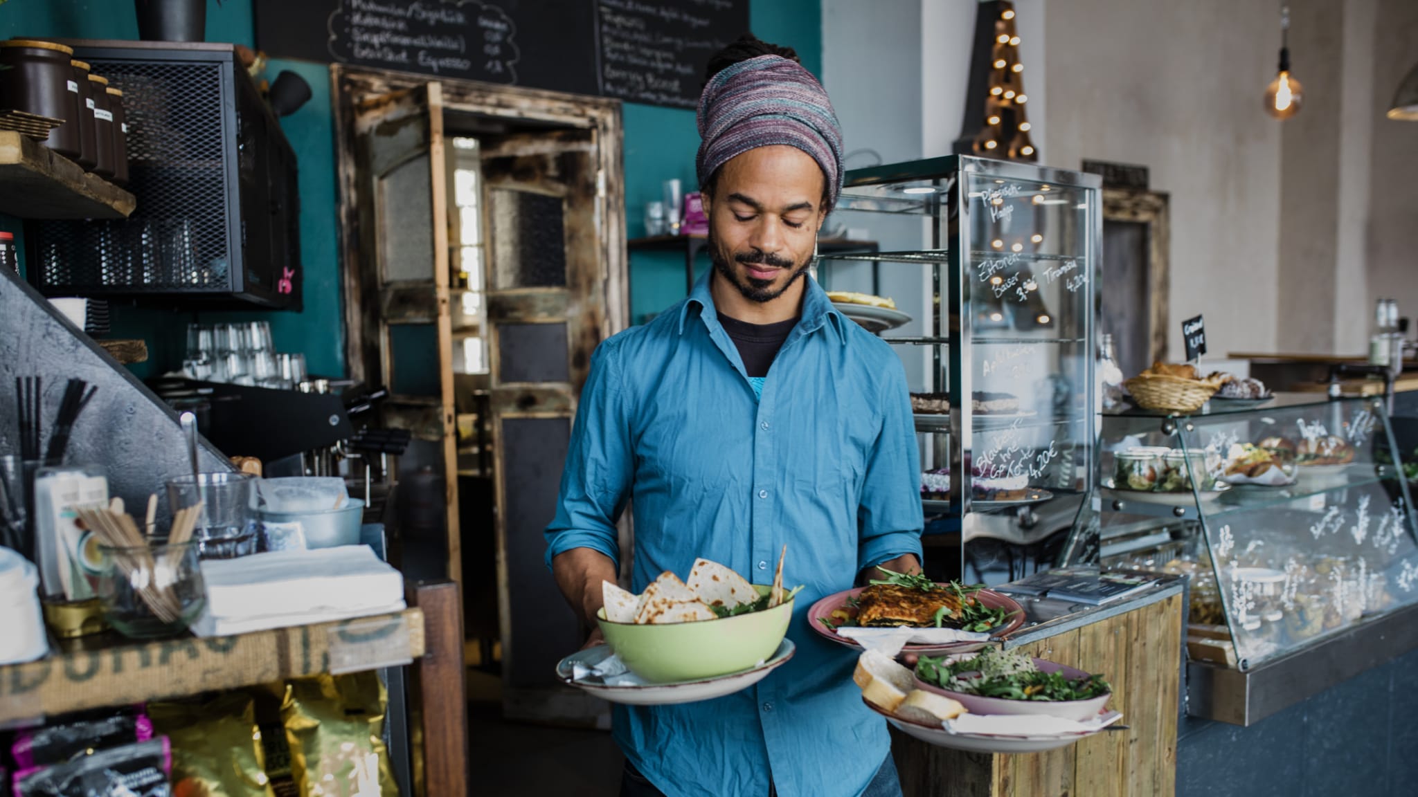 Ein Kellner trägt Essen zu einigen KundInnen während der Mittagszeit in einem veganen Café. © Hinterhaus Productions/DigitalVision via Getty Images