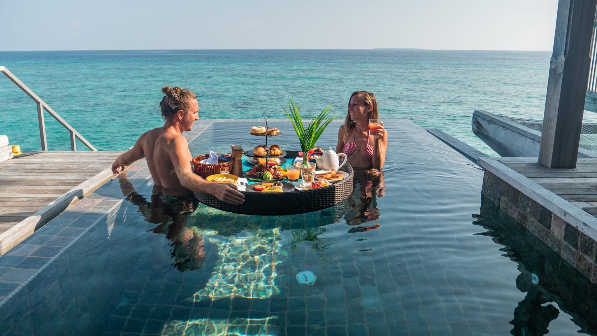 Ein Paar mit einem schwimmenden Frühstückskorb in einem Infinity Pool.