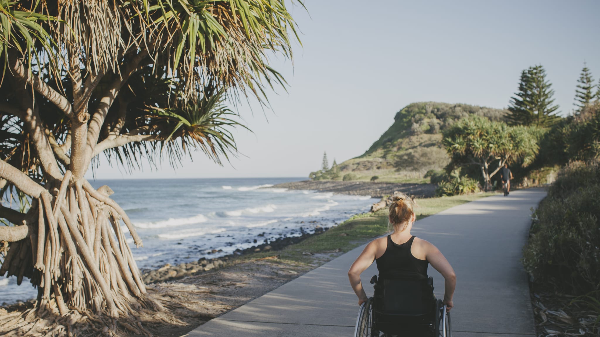 Eine Frau fährt in einem Rollstuhl auf einem Weg direkt am Meer.