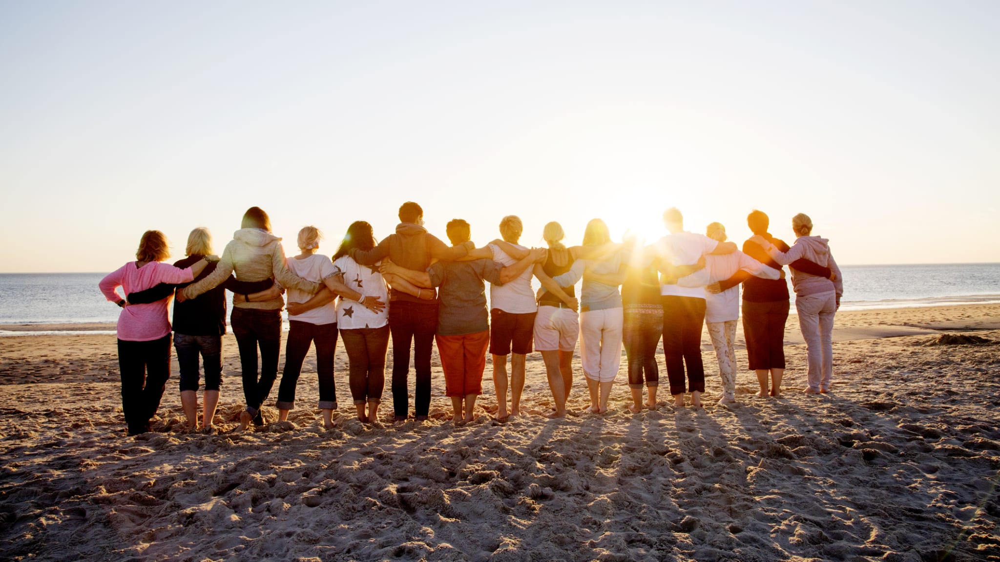Eine Gruppe Menschen schaut mit den Armen umeinander von einem Strand aufs Meer.