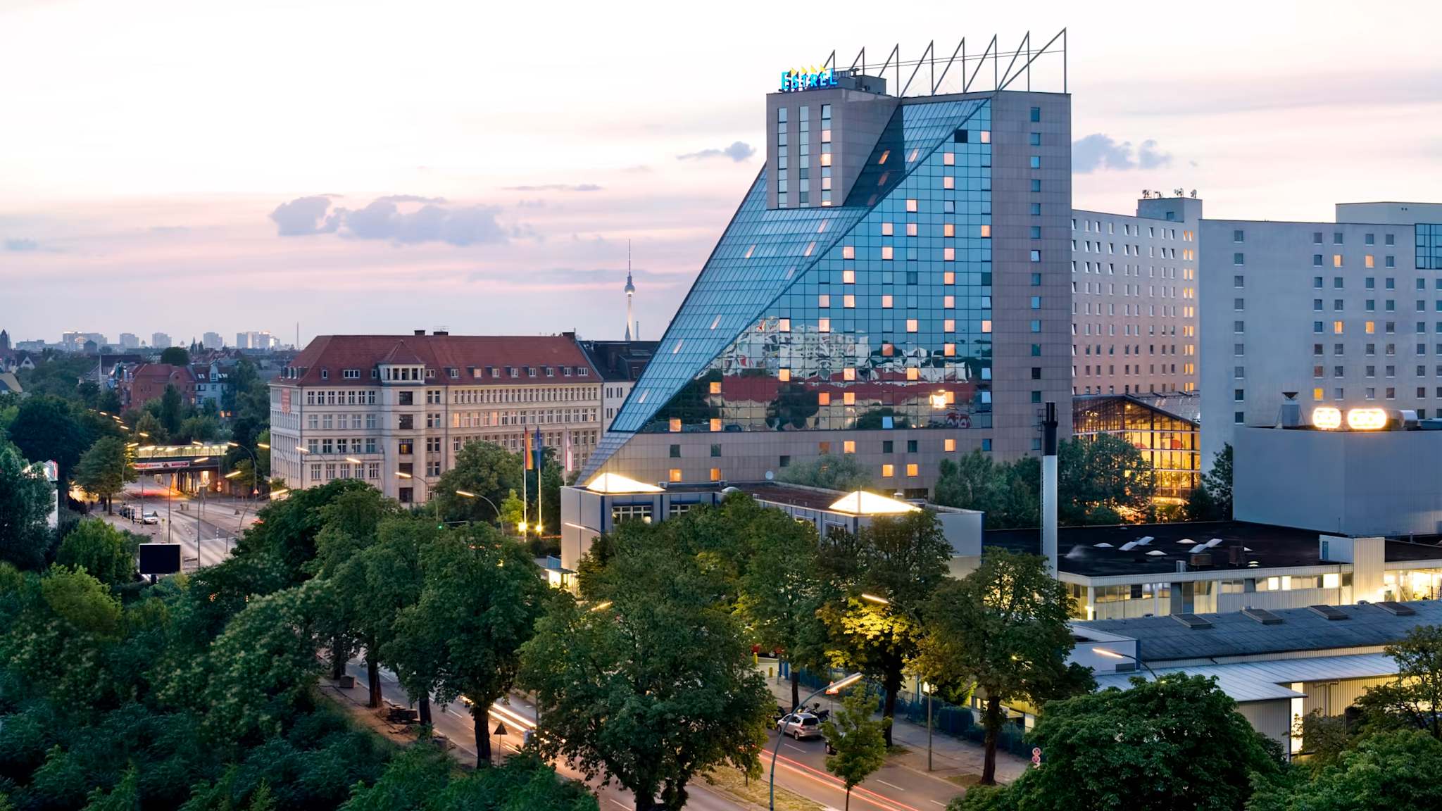 Außenansicht des Estrel Hotels in Berlin mit großer Glasfront und einer Allee davor bei Sonnenuntergang
