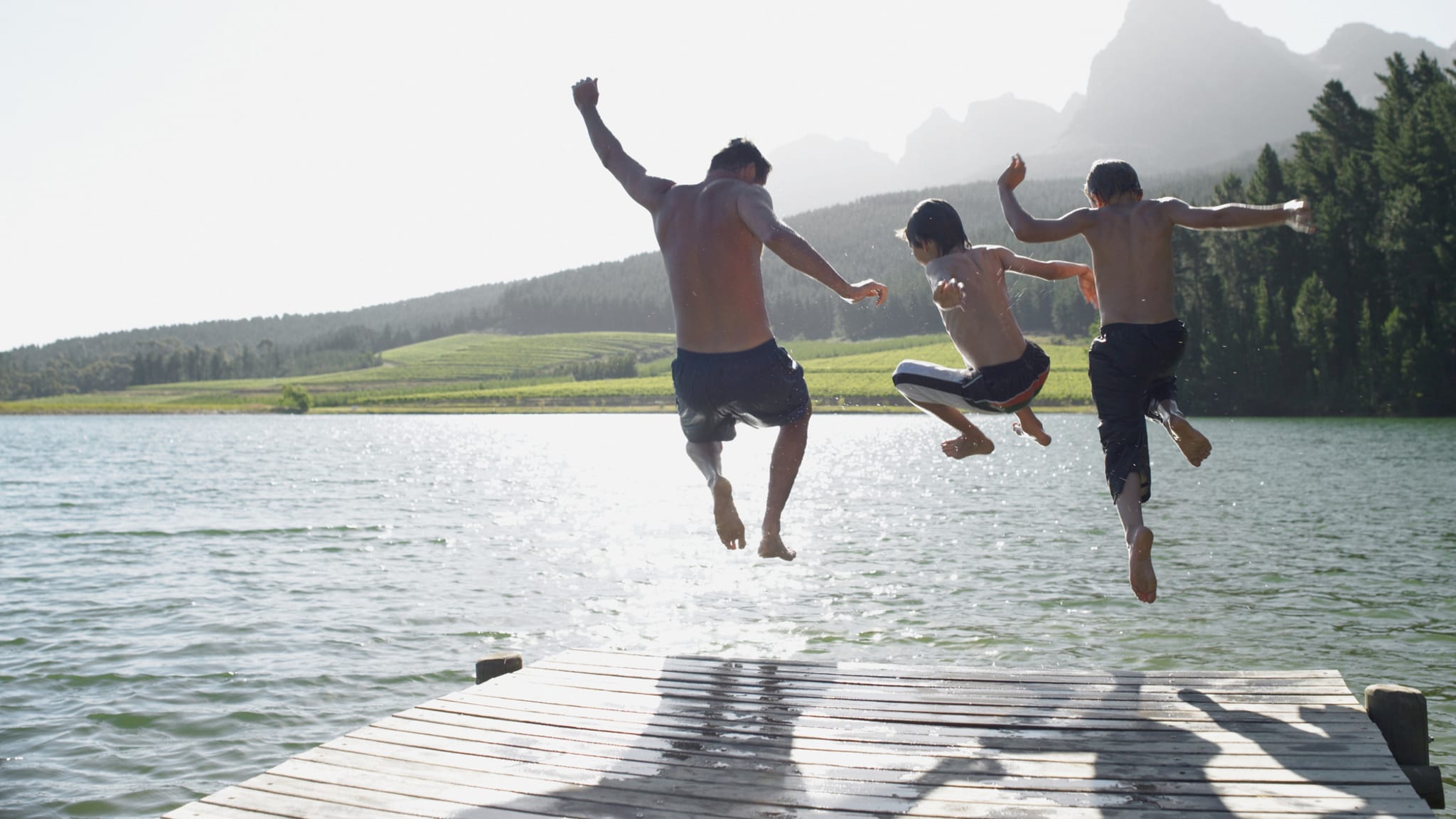 Ein erwachsender Mann und zwei Jungen springen in einen See von einem Steg aus an einem sonnigen Tag.
