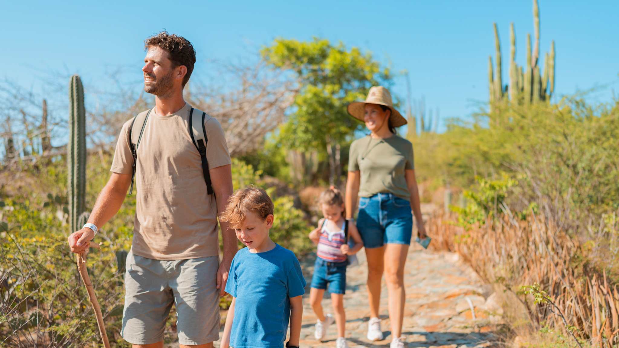 Eine Familie wandert einen Wanderweg entlang. Ein kleiner Junge läuft neben einem Mann mit Wanderstab. Dahinter läuft ein kleines Mädchen neben einer Frau mit Sonnenhut.