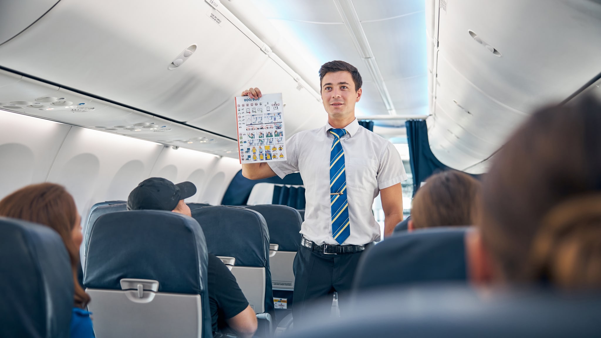 Ein Flugbegleiter in einem weißen Hemd und gestreifter Krawatte hält die Sicherheitsanweisungen hoch