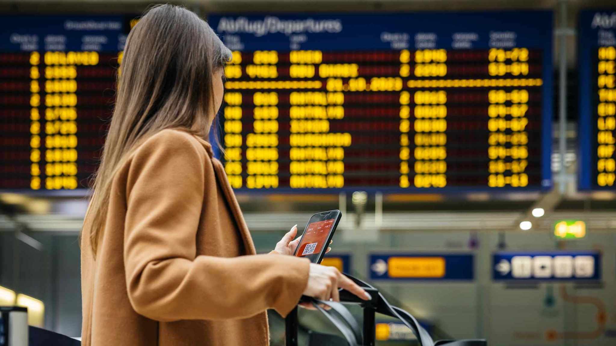 Frau mit Koffer und Handy in der Hand vor einer Anzeigetafel am Flughafen.