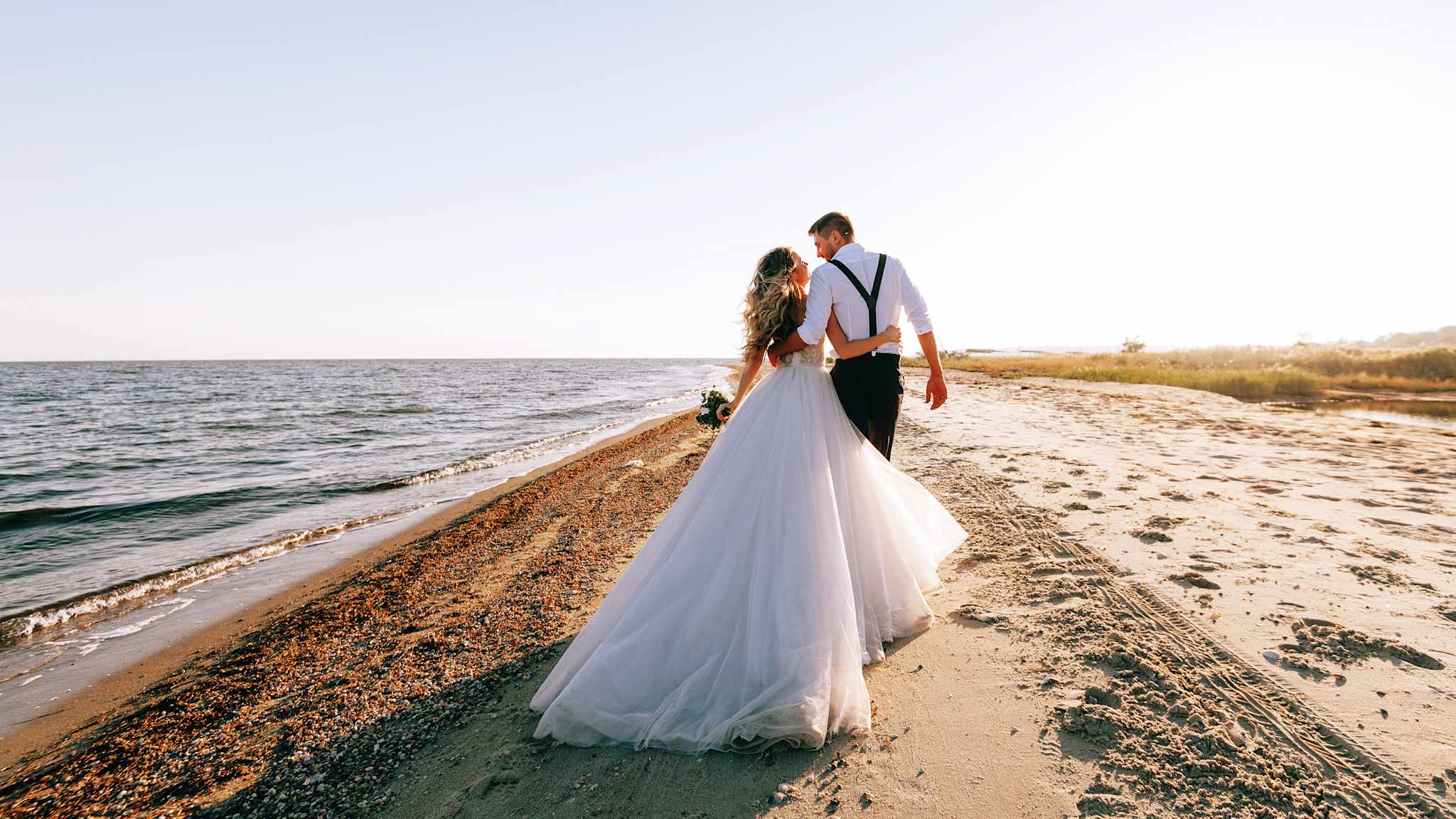 Eine Braut und ein Bräutigam spazieren Arm in Arm am Strand.
