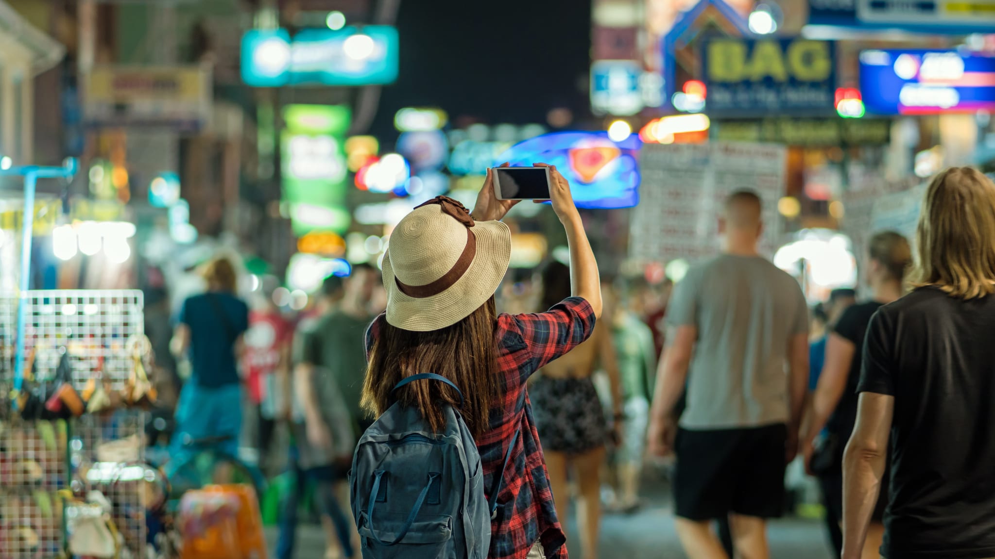 Eine Frau mit Hut, langen Haaren, kariertem Hemd und Rucksack fotografiert mit dem Smartphone auf einer Straße mit zahlreichen Neonschildern.