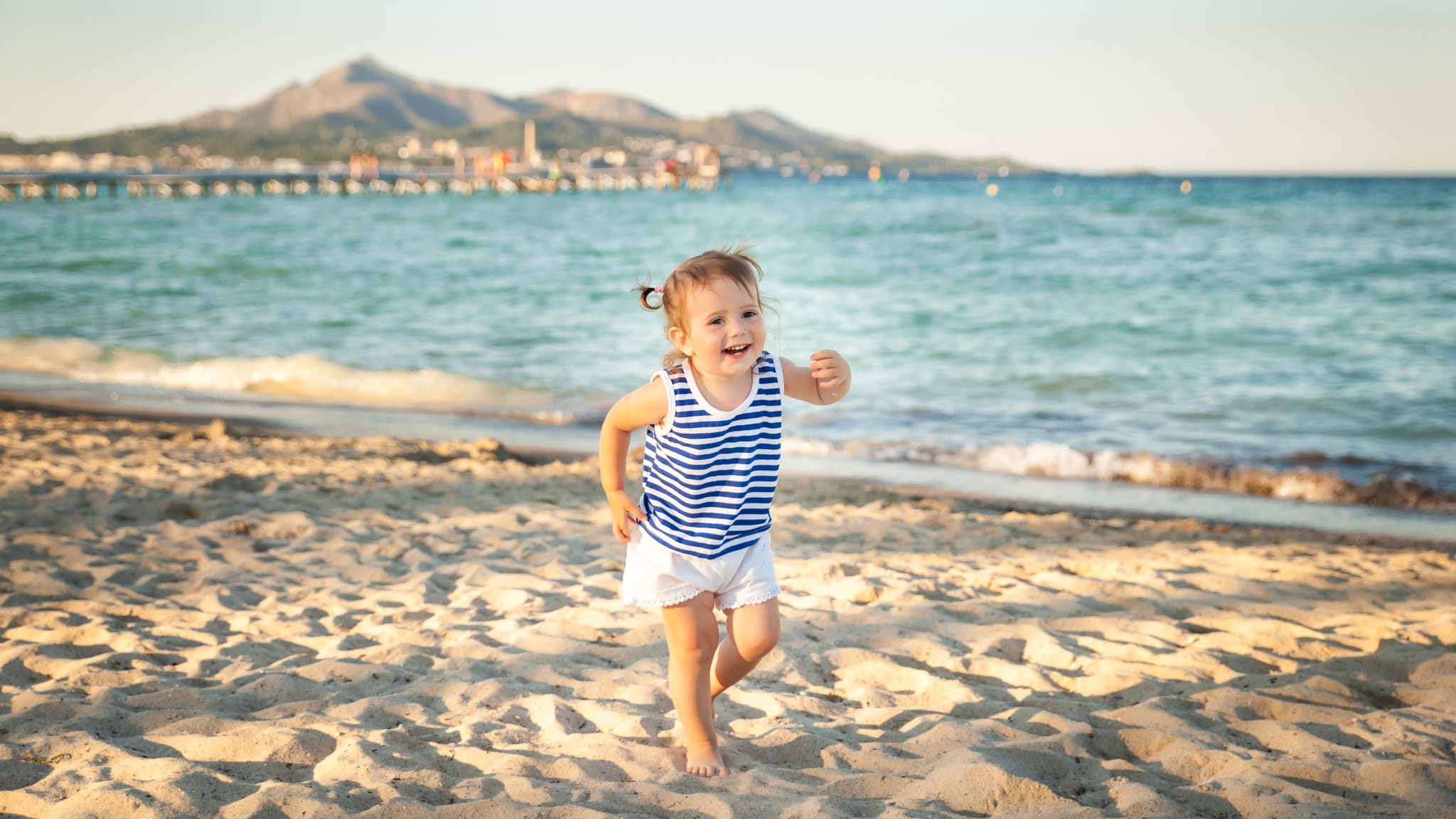 Kleines Mädchen in blau geringeltem Oberteil rennt am Strand von Mallorca, Spanien