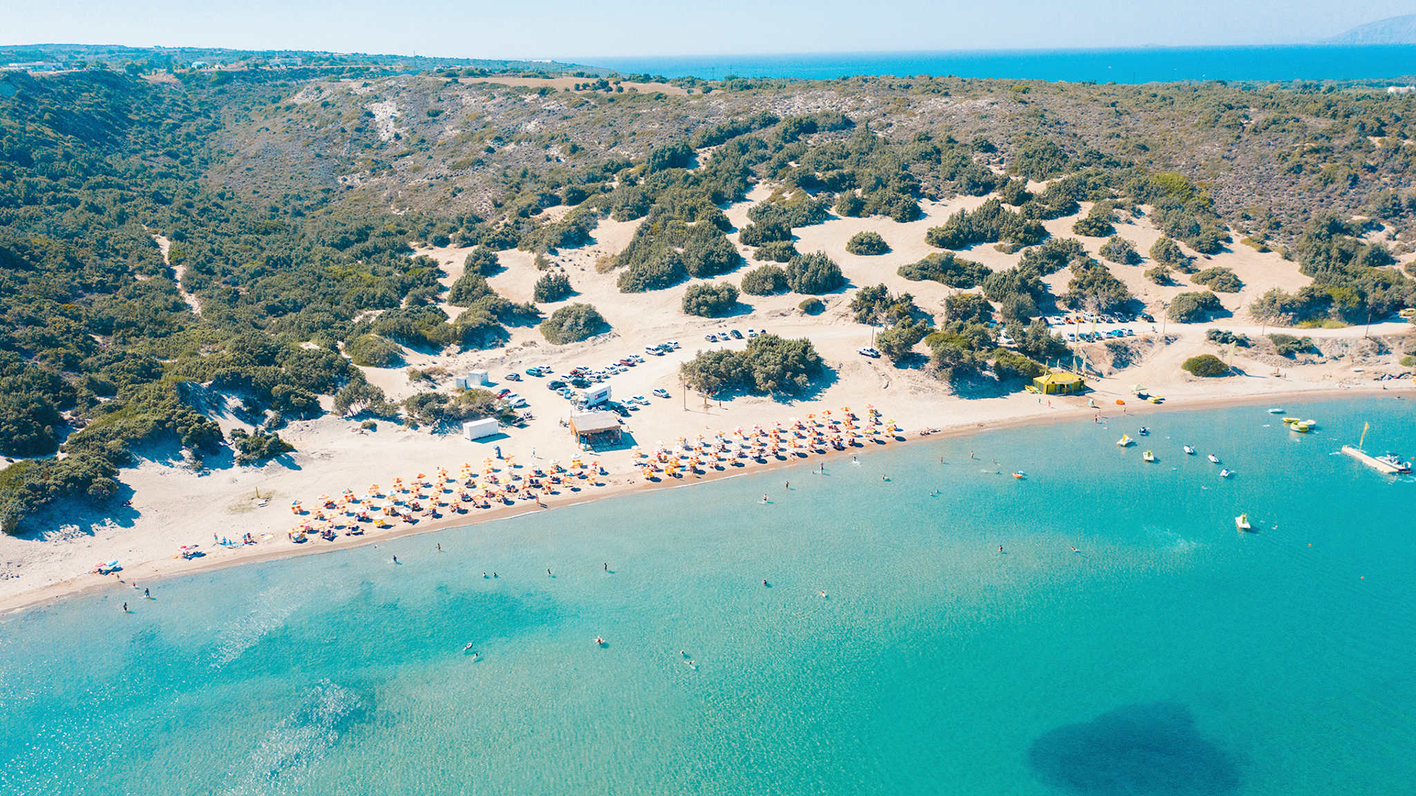 Ein weißer Sandstrand mit Sonnenschirmen grenzt an das klare Meer. Im Hintergrund sind viele Sträucher zu sehen, die auf Dünen wachsen.