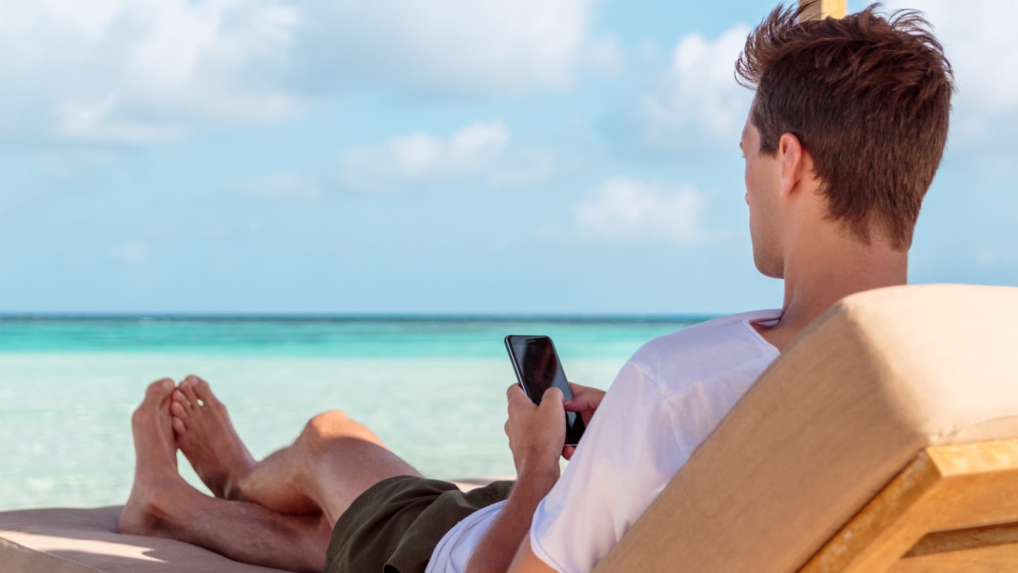 Mann liegt mit Handy in der Hand auf einem Liegestuhl am Strand.