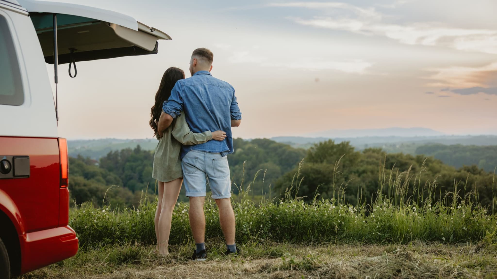 Ein Paar steht vor einem Van und schaut Arm in Arm in die Natur hinaus.