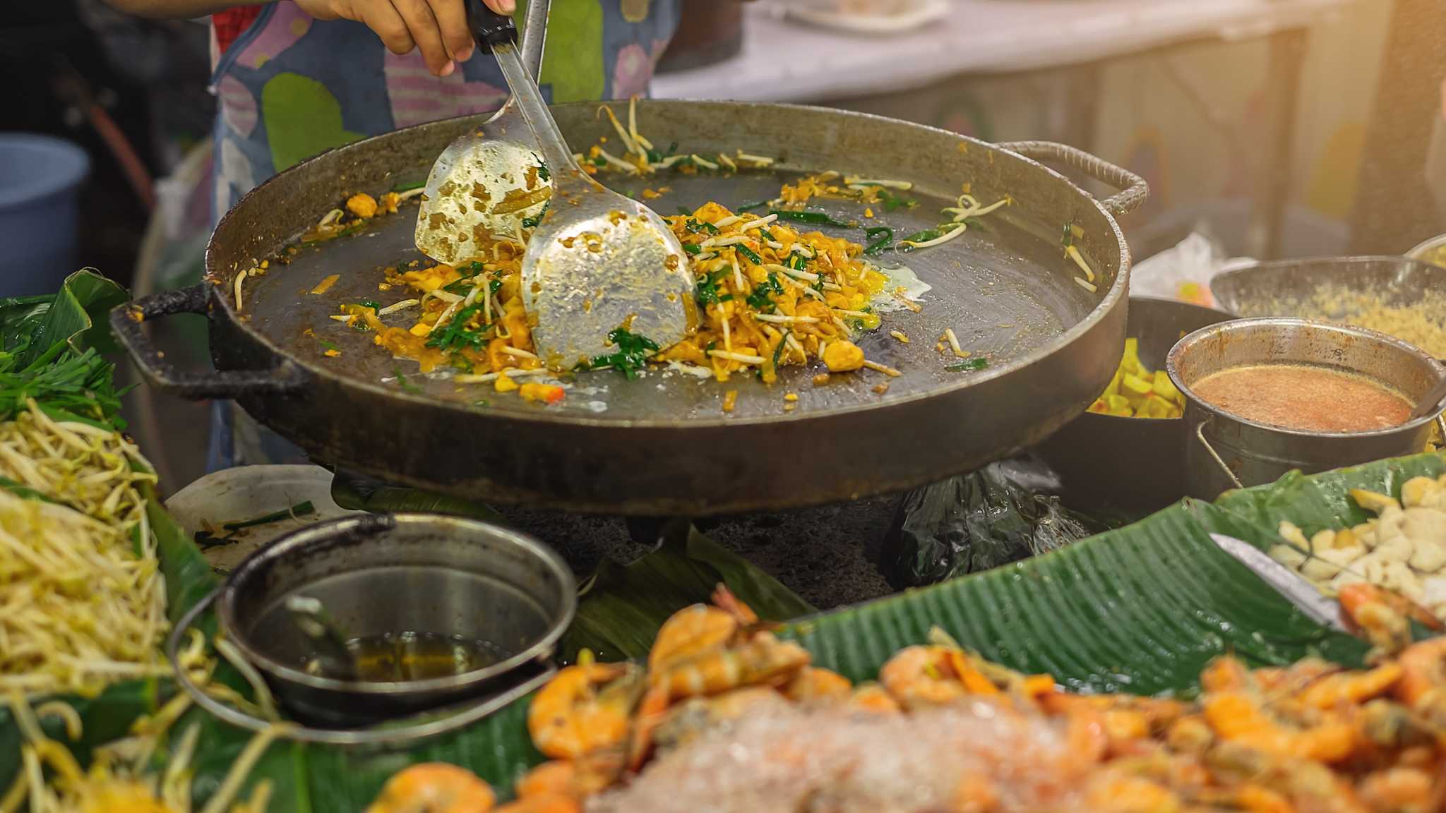 Eine Frau bereitet Pad Thai auf einem Markt in einer großen Pfanne zu.