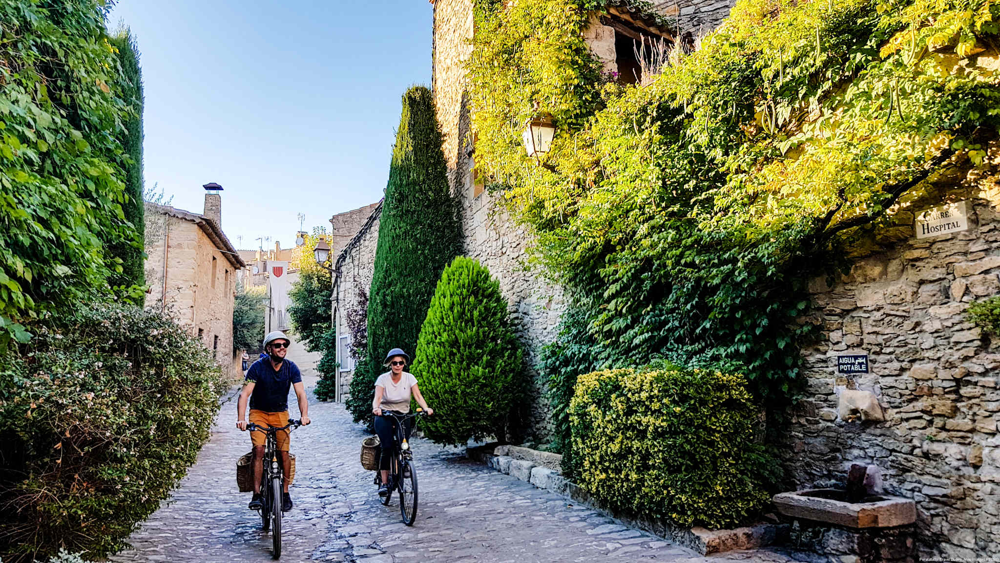 Ein Paar fährt mit einer Burricleta (Elektrofahrrad) eine gepflasterte Straße in Peratallada entlang.