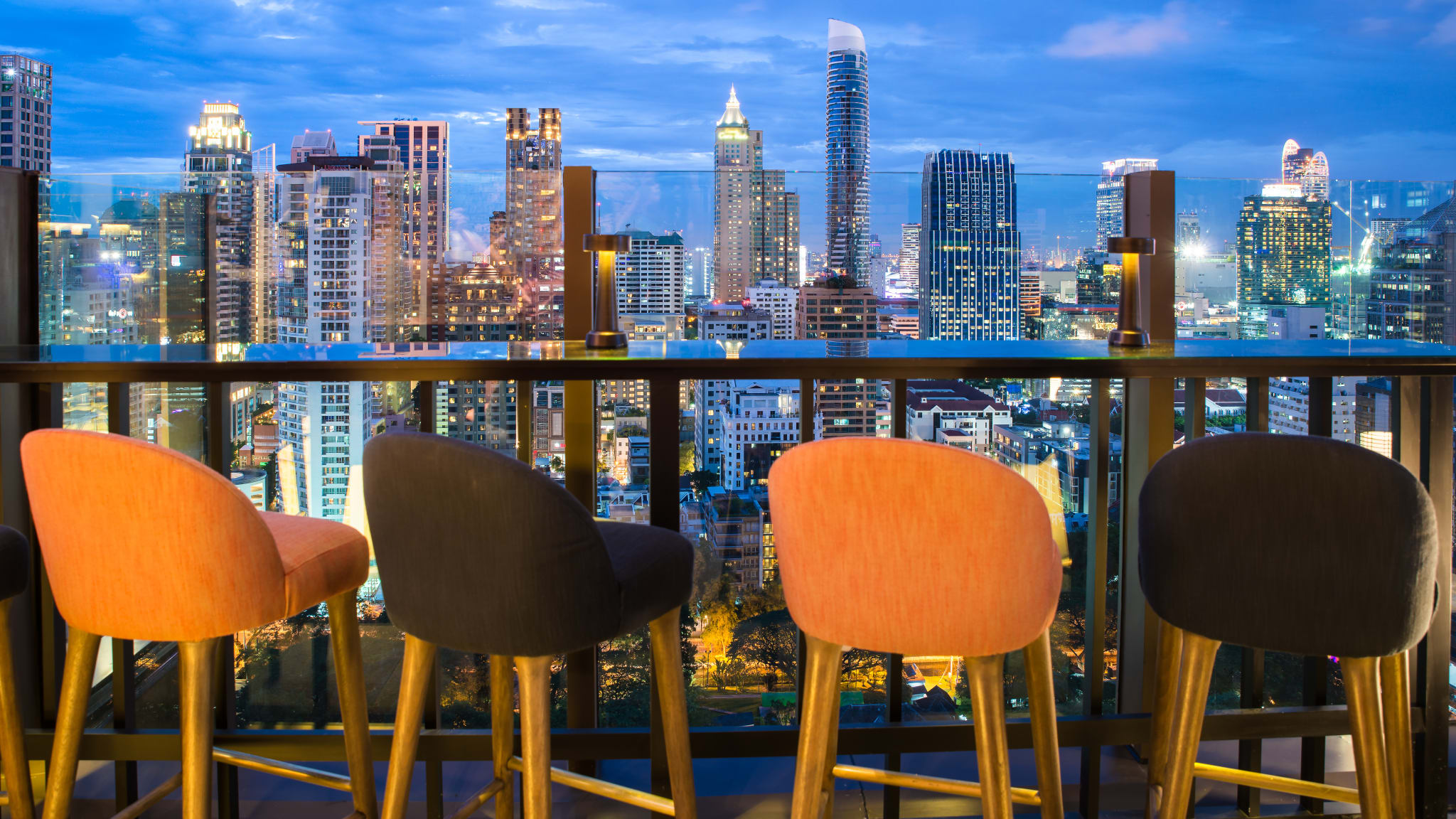 Vier Barstühle stehen an einem Tisch an einer Brüstung mit Blick auf die Hochhäuser von Bangkok.