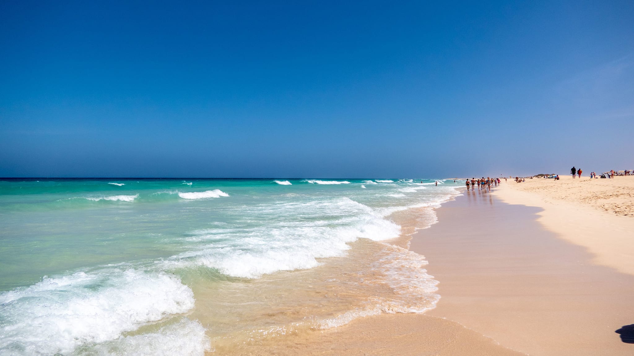 Ein langer hellgelber Sandstrand, hellblaues Meer, in der Ferne spazieren Menschen auf Fuerteventura