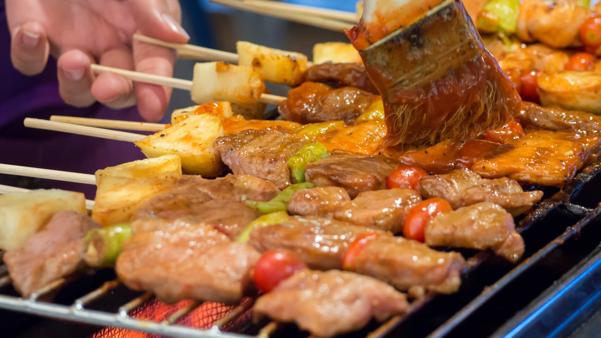 Spieße mit Fleisch und Gemüse liegen auf einem Grill und werden mit einer Satay Soße bestrichen