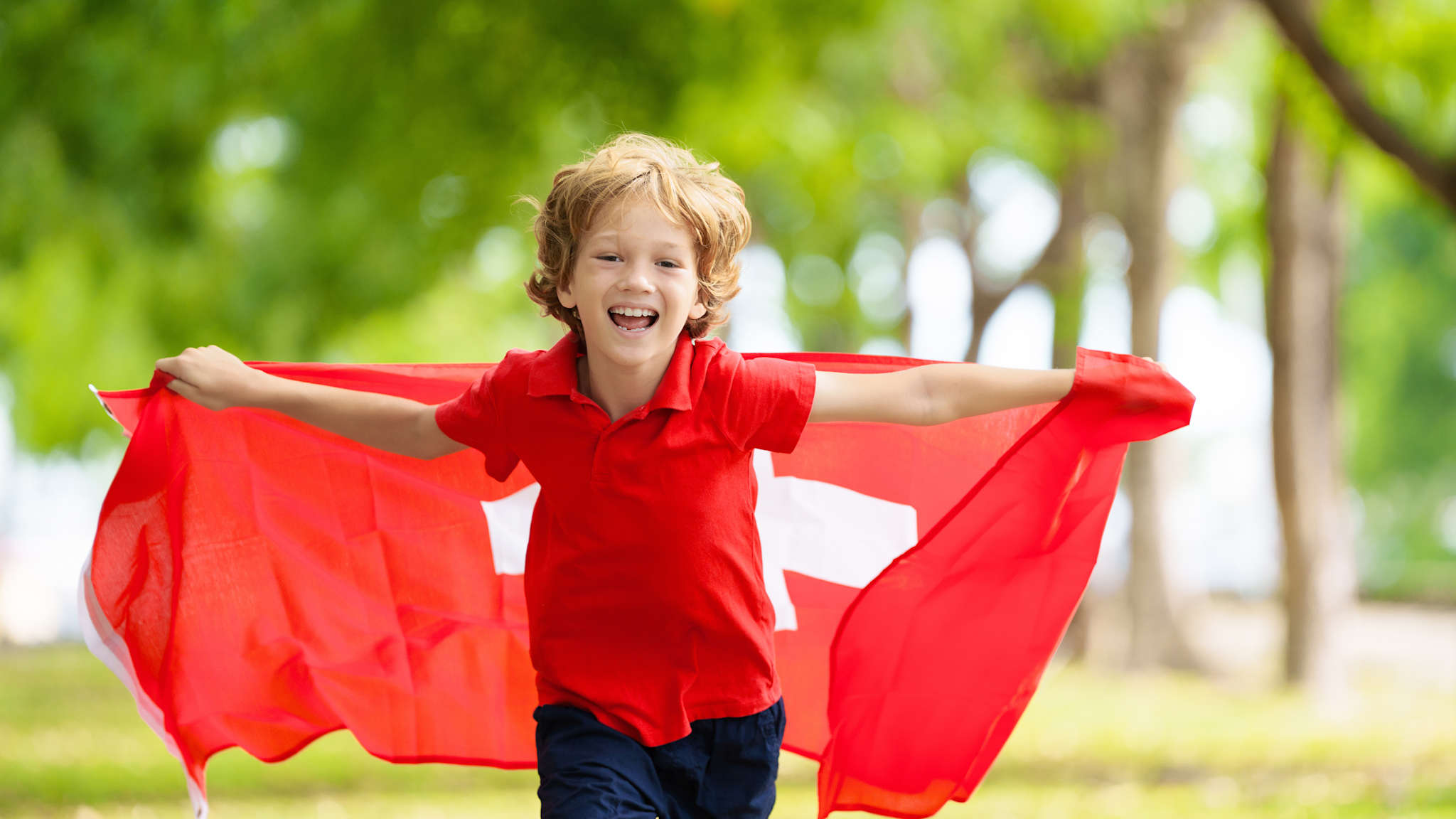 Ein Junge läuft mit einer großen Schweiz Flagge durch einen Park und lacht