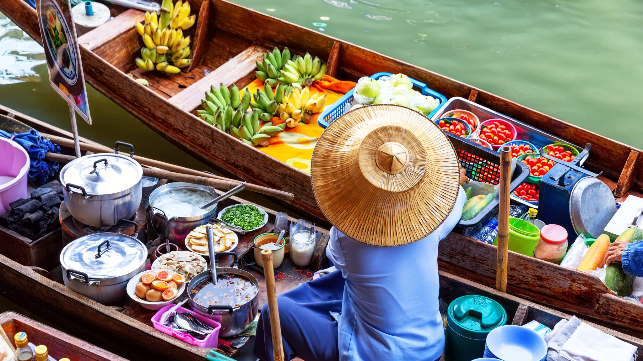 Ein Mann mit traditionellem thailändischem Strohhut sitzt auf einem Boot. Um ihn herum stapeln sich Töpfe und Teller. In einem zweiten Boot neben ihm liegen Schalen mit Tomaten und Bananen.