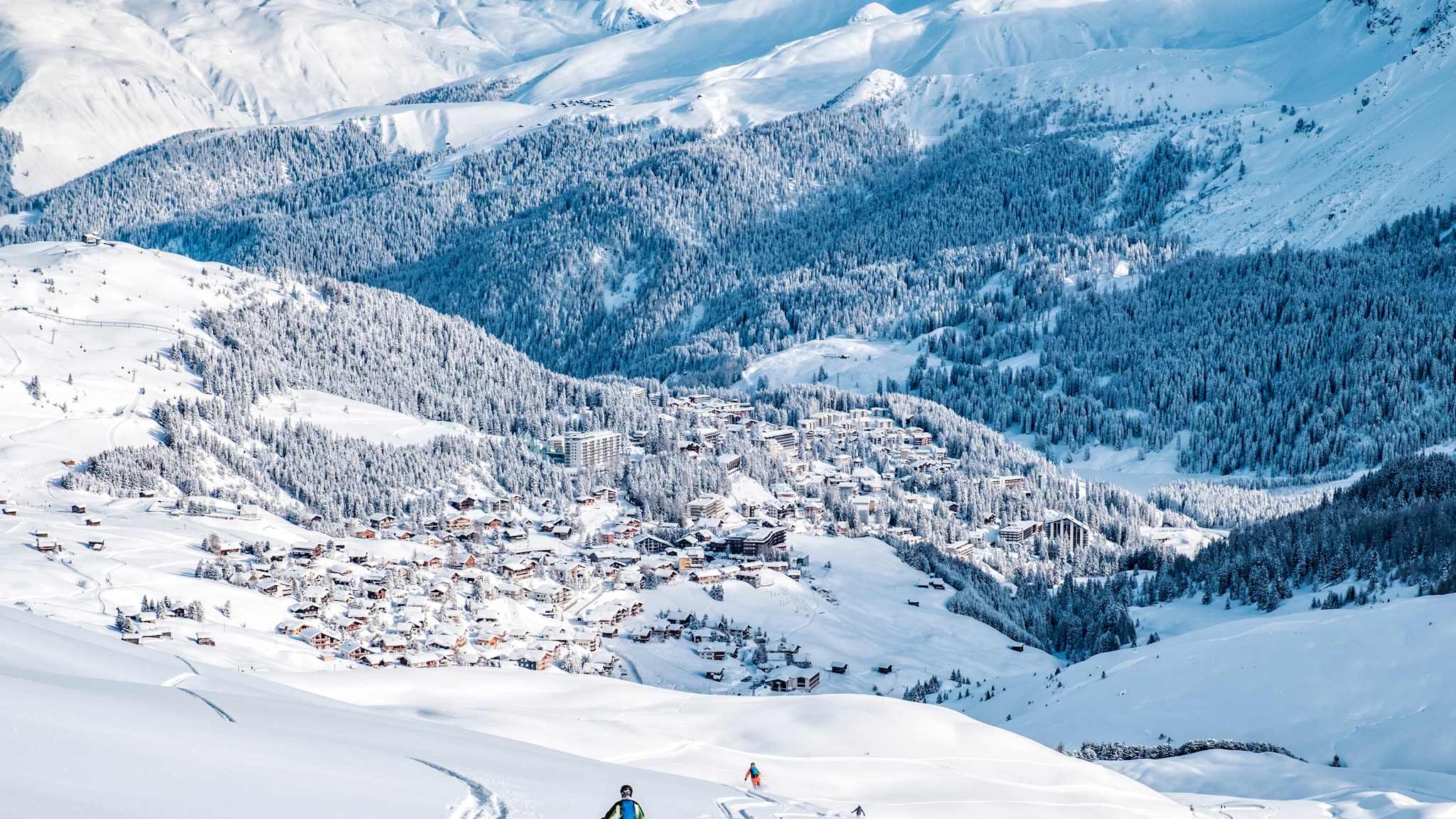 Skifahrer fahren einen Abhang hinunter, mit Blick auf eine eingeschneite Stadt in den Bergen.